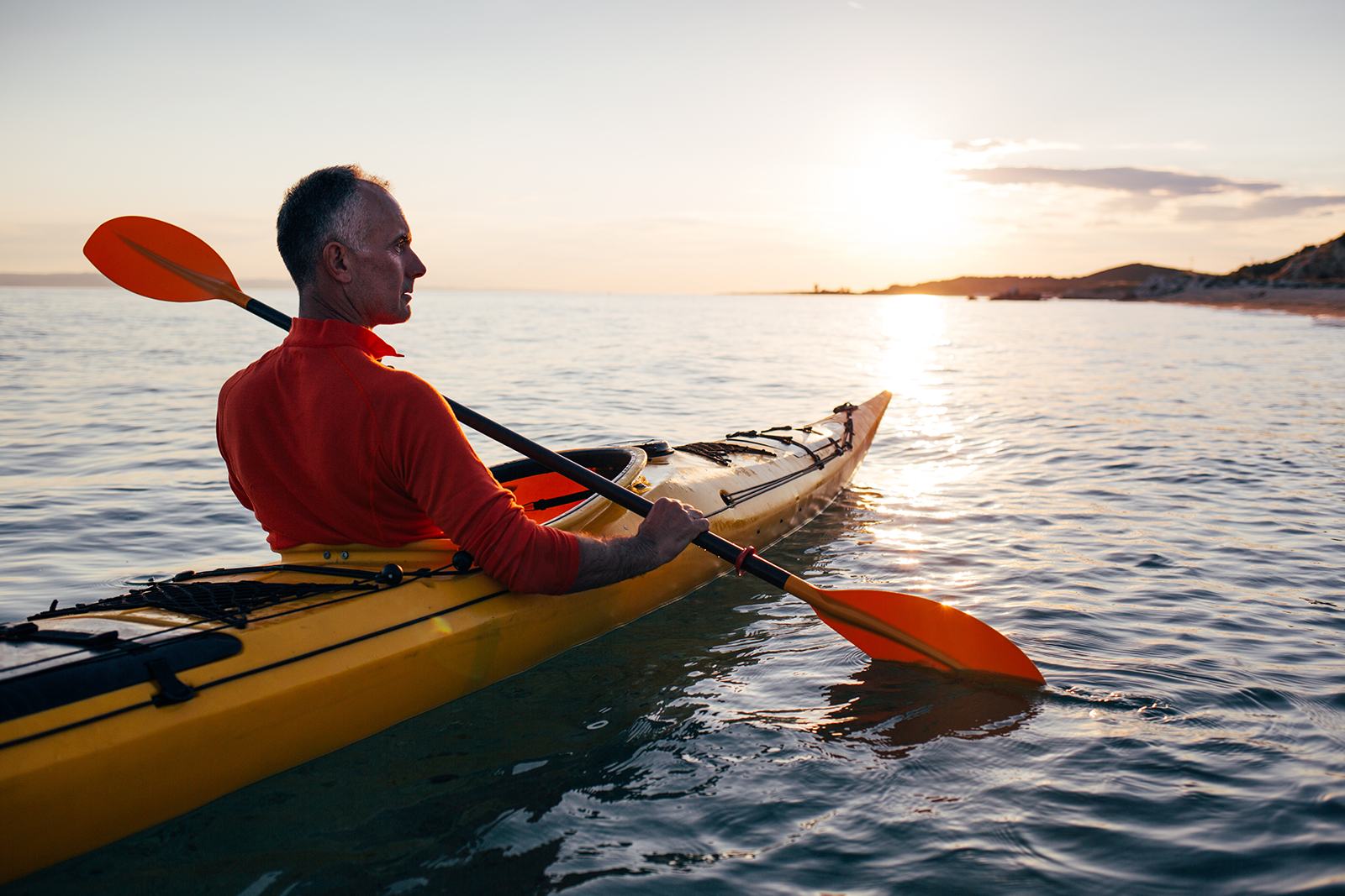 Man in a red long-sleeve shirt paddling a yellow kayak at sunset on calm water.