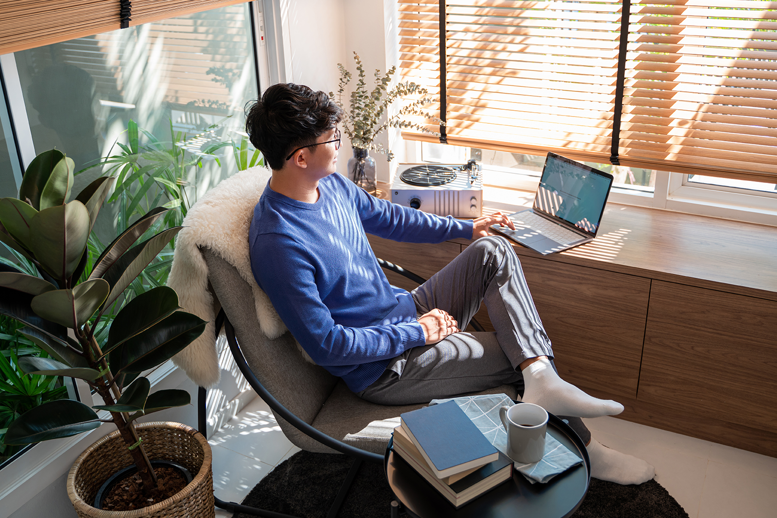 Man in a blue sweater sits in a cozy home office, working on a laptop at a wooden desk with plants nearby.