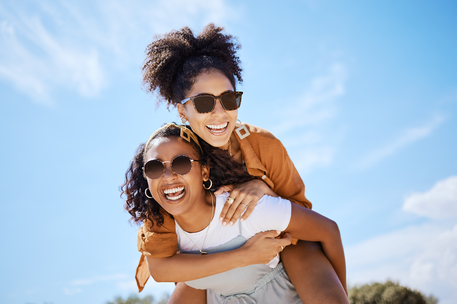 Two women laughing outdoors; one gives the other a piggyback ride under a bright blue sky, both wearing sunglasses.