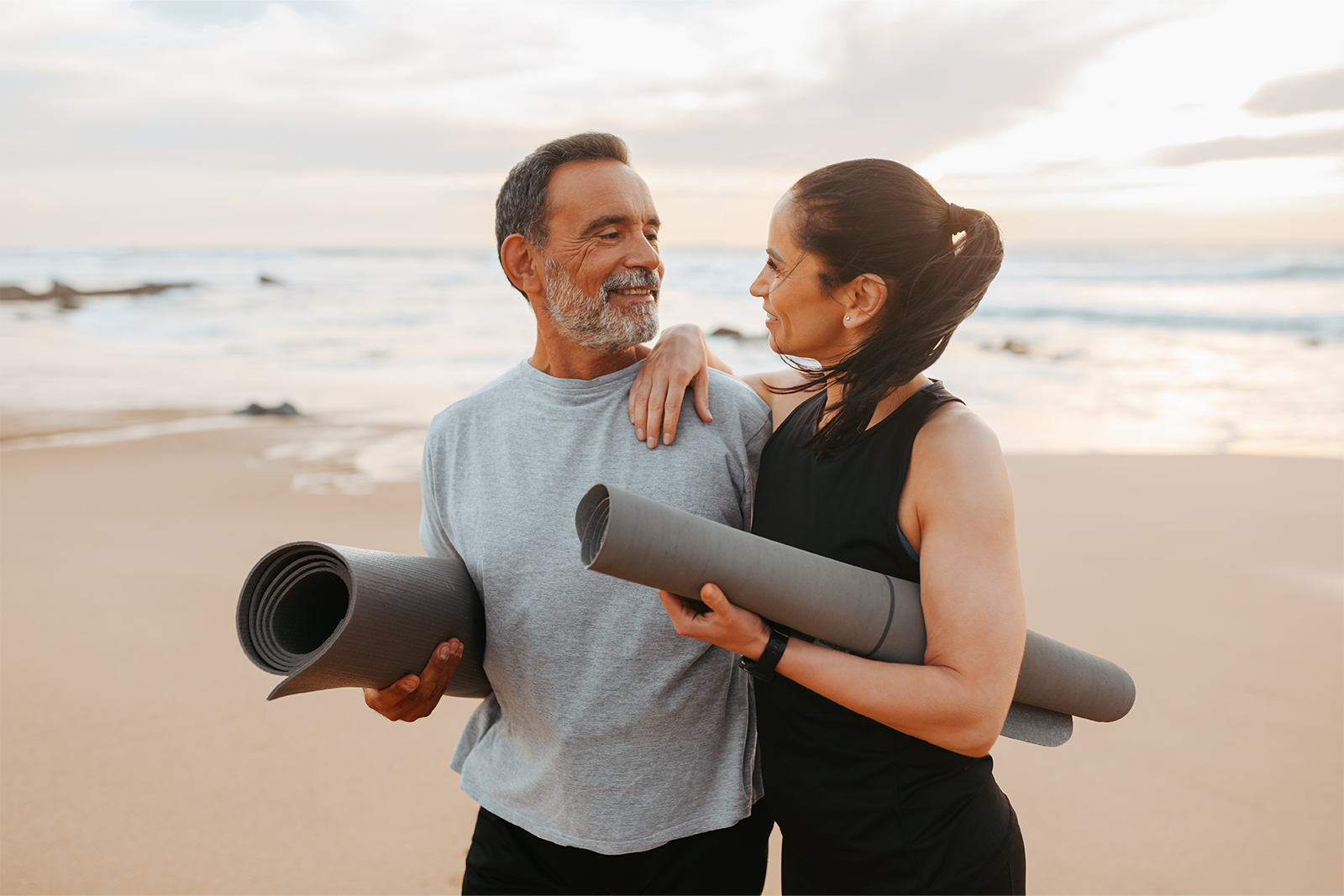 Couple on a beach at sunset carrying rolled yoga mats, smiling at each other while preparing for a workout.